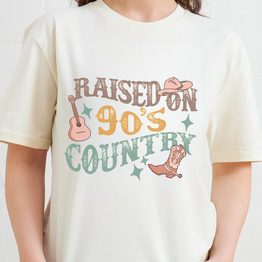 White t-shirt with 'Raised on 90's Country' graphic design featuring a guitar, cowboy hat, and boot.