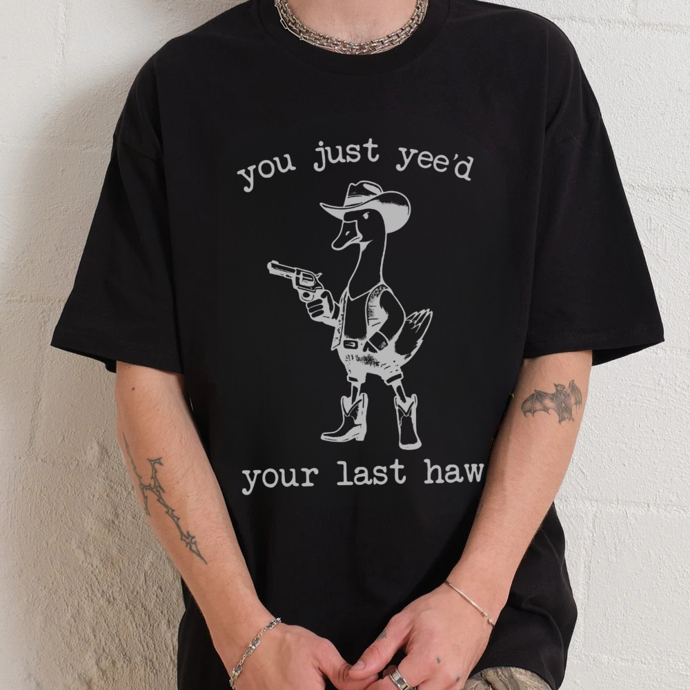Person wearing a black t-shirt with a graphic and text, sitting against a white wall.