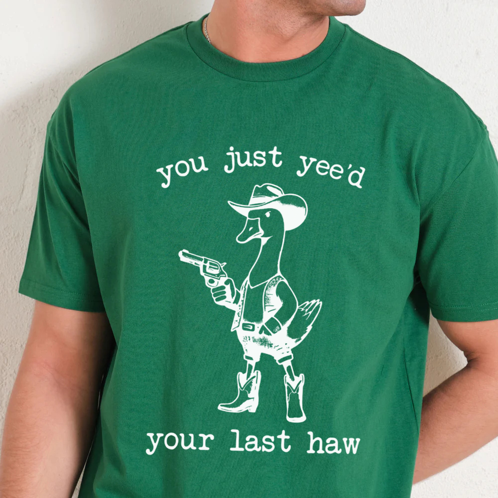 Green t-shirt with a graphic of a duck in a cowboy hat and boots, holding a gun, with text on a plain background.