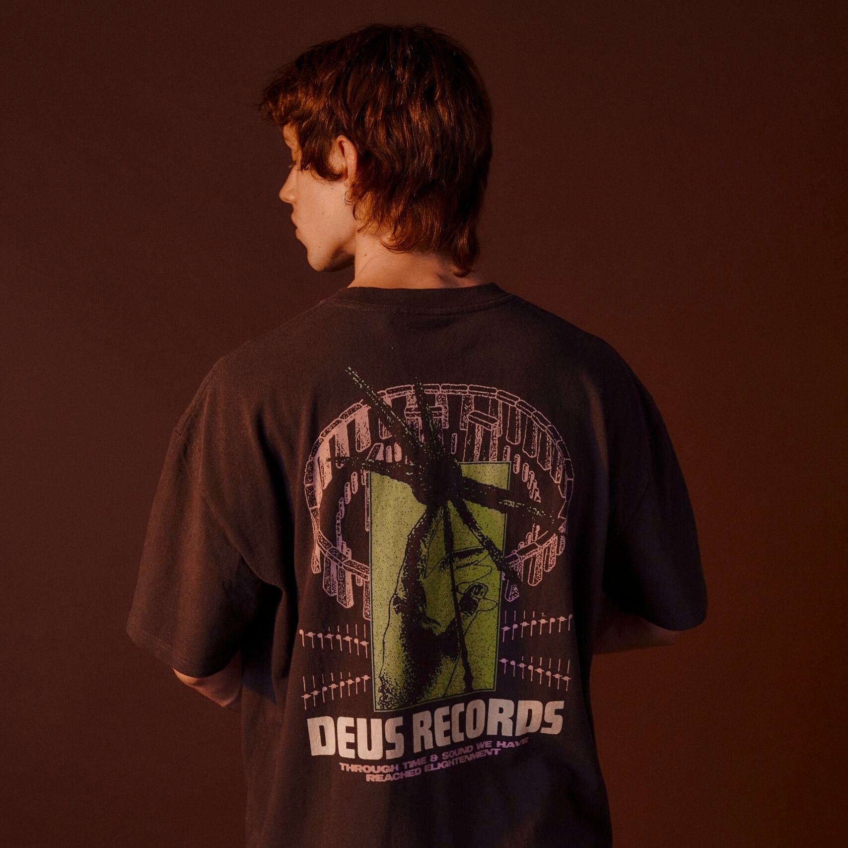 Person wearing a black t-shirt with 'Deus Records' graphic on the back against a brown background