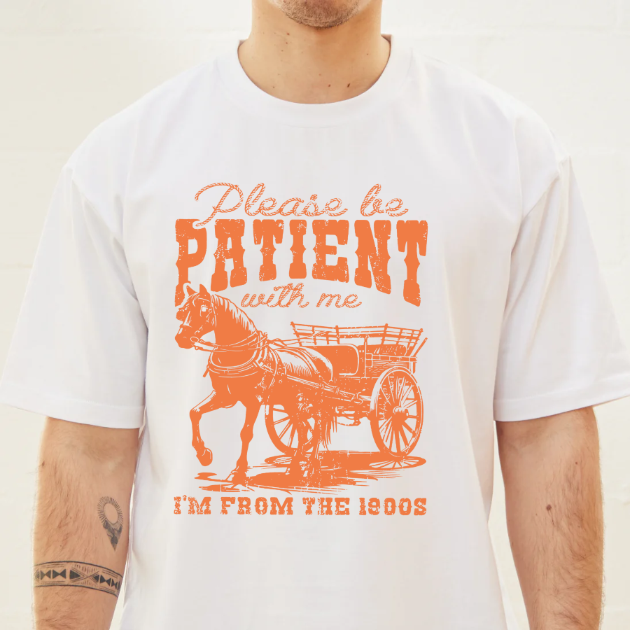 White t-shirt with orange graphic and text 'Please be patient with me, I'm from the 1900s' on a plain background
