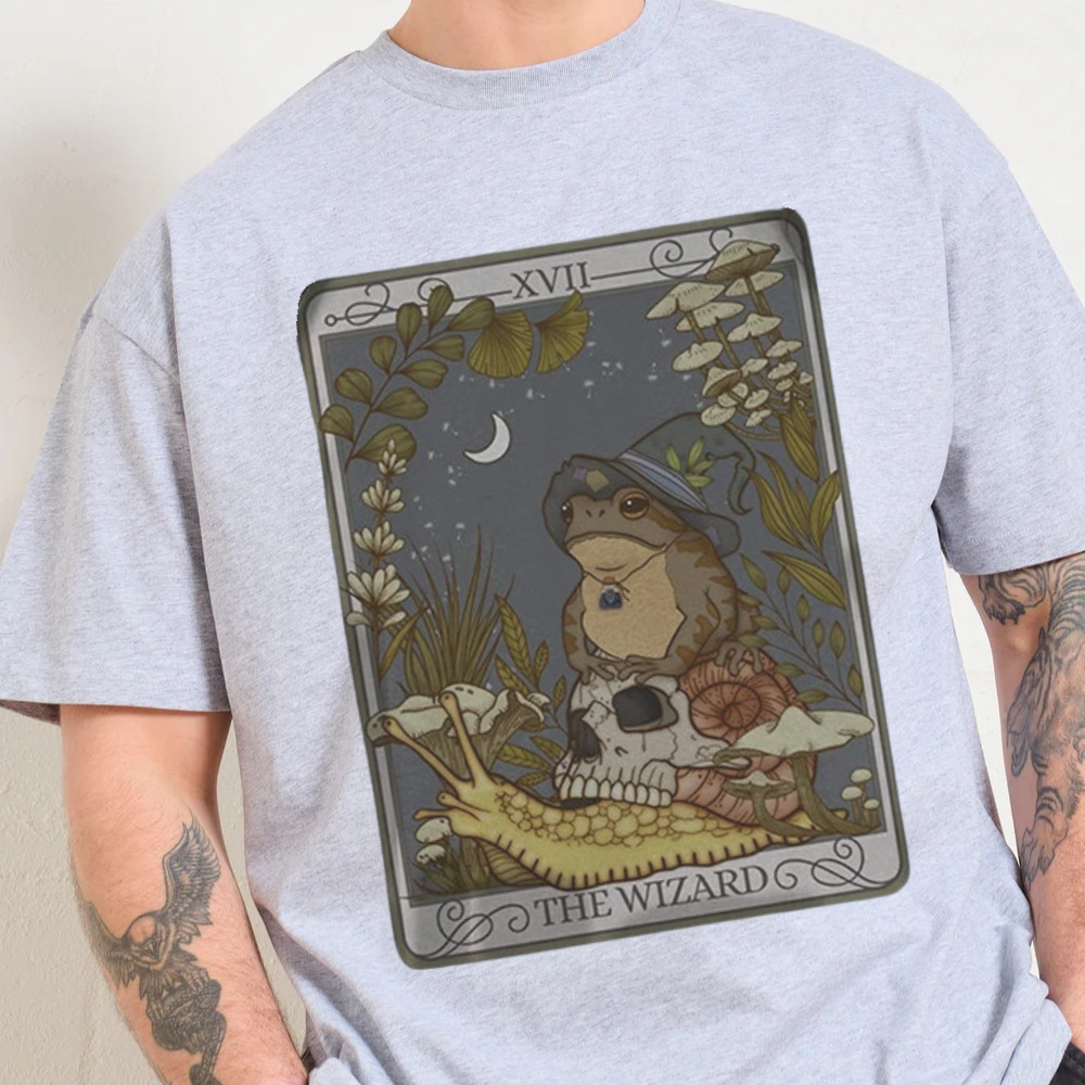 Person wearing a gray t-shirt with a graphic of a frog in a wizard hat and scarf, sitting on a skull with plants and a moon in the background.
