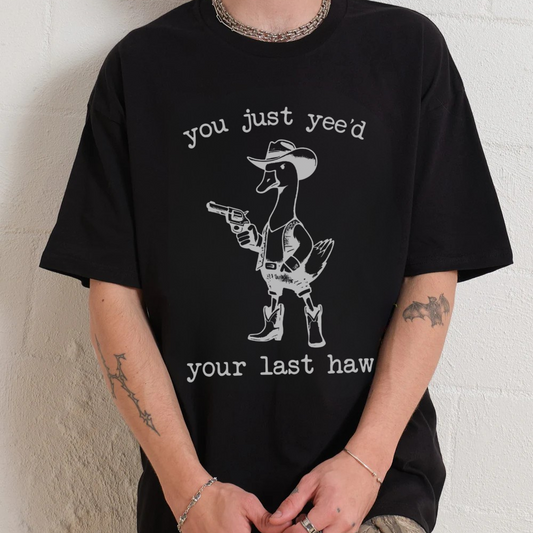 Person wearing a black t-shirt with a graphic and text, sitting against a white wall.